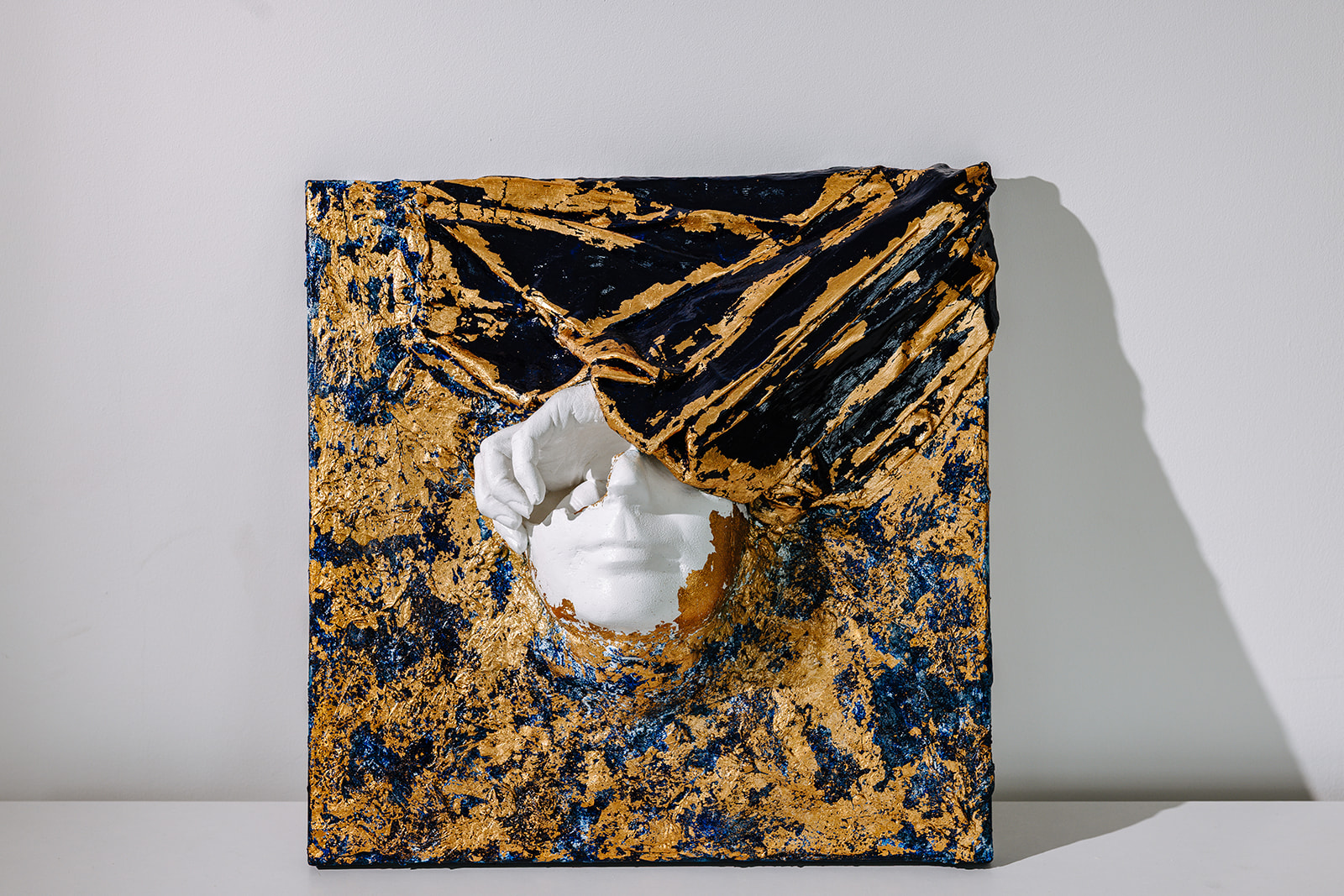 Hochrelief Kunstwerk 2024 von Marianna Ackermann, Mixed Media Wandobjekt mit Gips, Stoff und Blattmetall, plastische weiße Form auf Blau-Gold-Struktur, 40x40 cm